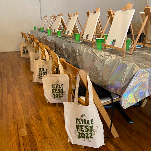 <p>Table and chairs set up for painting with tote bags hanging off the chairs</p>
