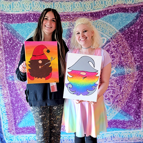 Two women smiling and holding paintings in front of a tapestry