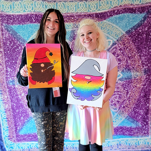 <p>Two women smiling and holding paintings in front of a tapestry</p>
