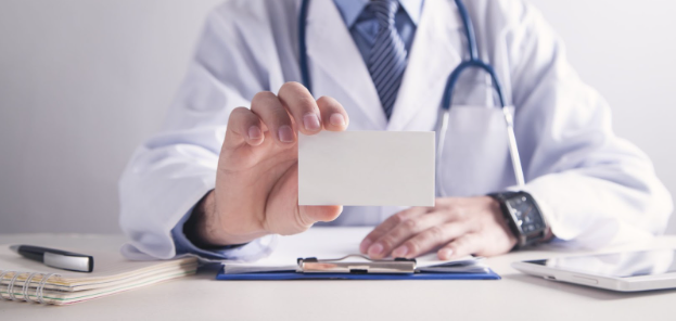 A doctor holds up a card while sitting at a desk with a clipboard and notepad
