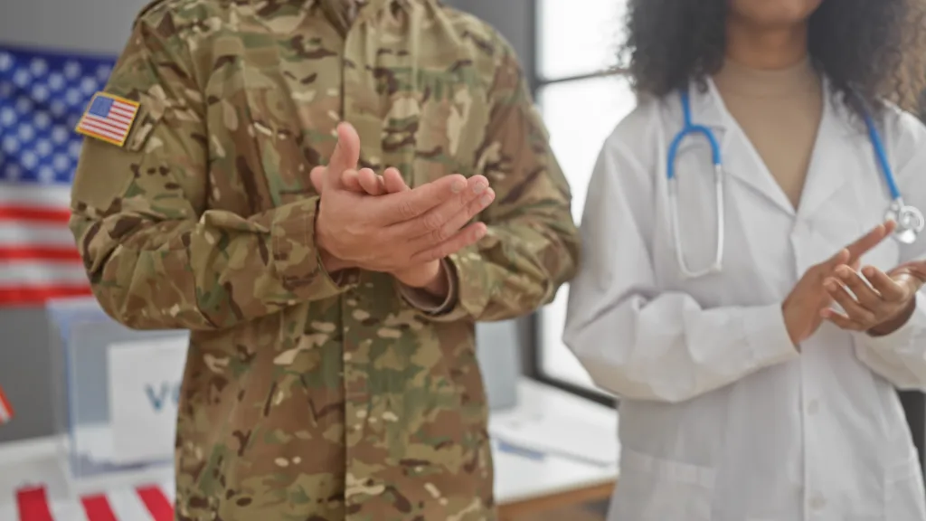 A person in military uniform and a person in a white medical coat with a stethoscope are clapping, with American flags visible in the background.