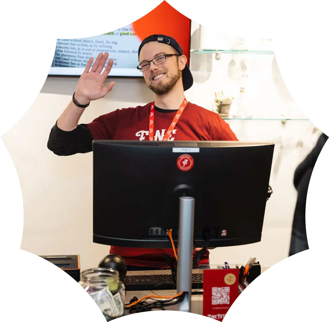 A smiling person wearing glasses and a cap waves while standing behind a computer monitor at a CT dispensary counter.