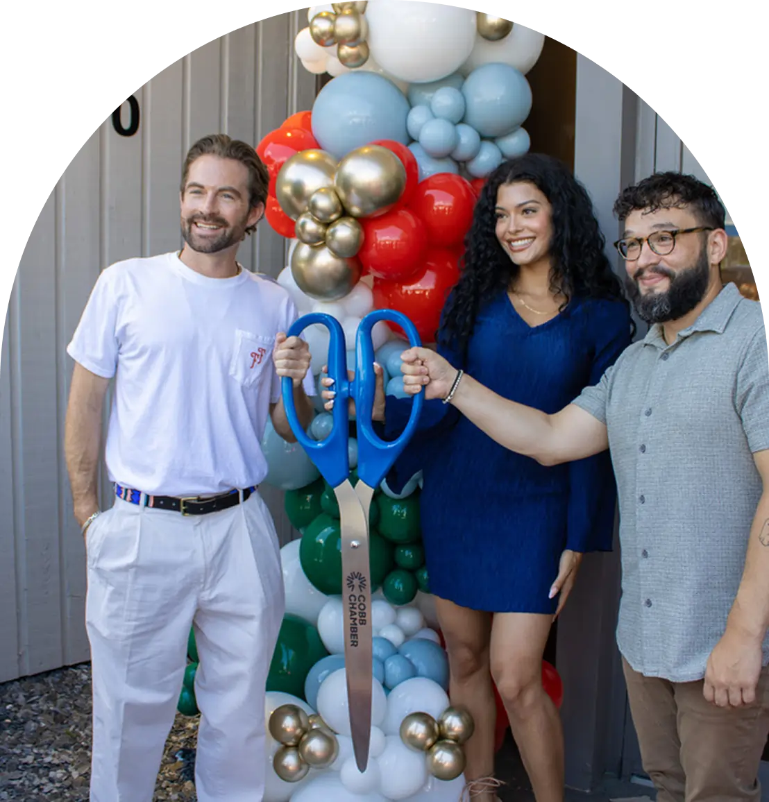 Three smiling people stand in front of a balloon arch at a Georgia dispensary, holding oversized blue scissors together as if for a ribbon-cutting ceremony.