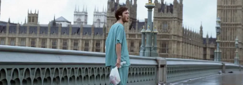 A man in hospital scrubs stands alone on an empty bridge in front of the Houses of Parliament in London, holding a plastic bag, with overcast skies above.