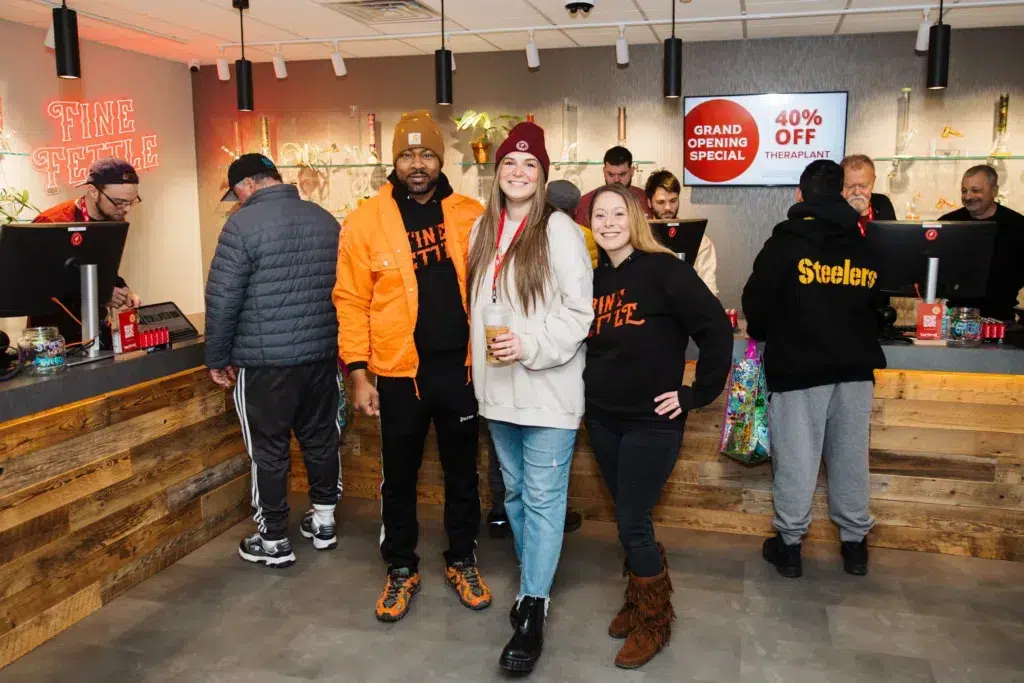 Three people pose and smile in front of a counter at a store; other customers and staff are in the background. Signs advertise a grand opening and a 40% off special.