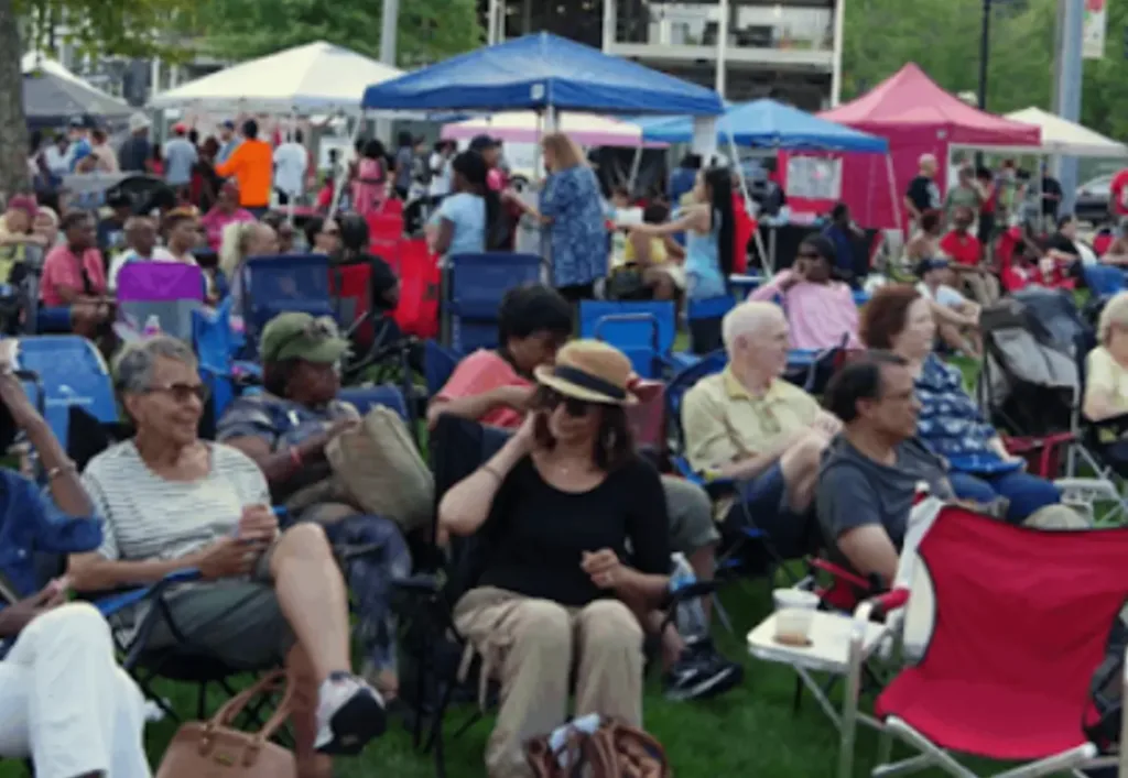A diverse crowd sits on lawn chairs at an outdoor festival, surrounded by tents and enjoying food and conversation on a grassy area.