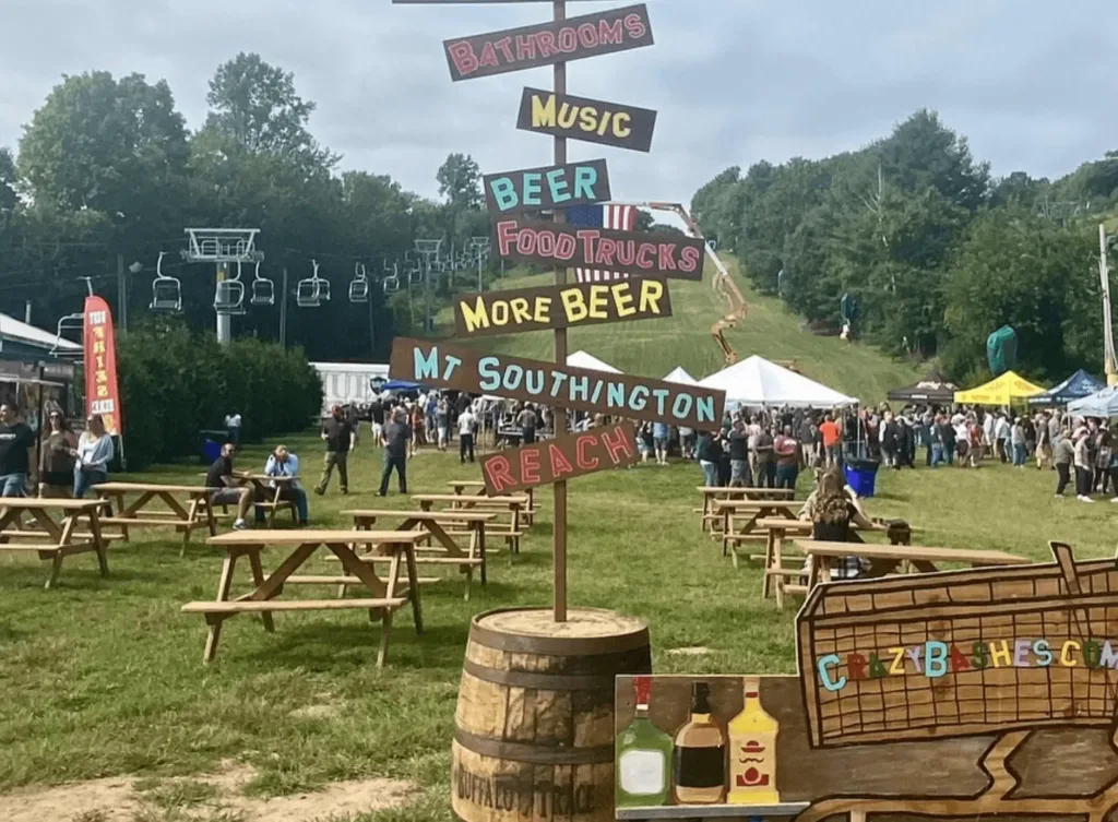 Outdoor festival scene with wooden signpost listing music, beer, food trucks, and other directions; picnic tables and crowds on a grassy area with tents and ski lifts in the background.
