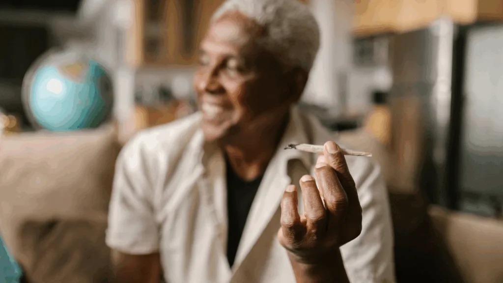 An older man with gray hair sits indoors, smiling and holding a lit joint in his hand. The background is softly blurred.