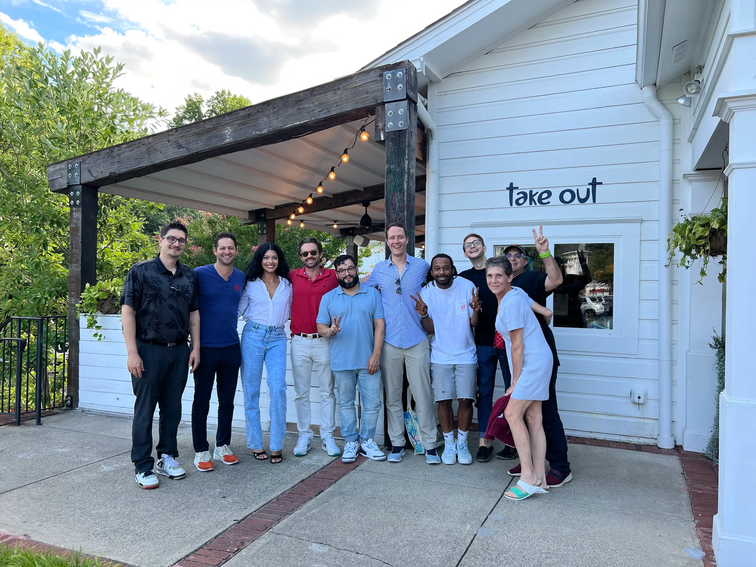 A group of eleven people smiling and posing outside a white building with a take out sign, greenery, string lights, and a georgia dispensary nearby on a sunny day.
