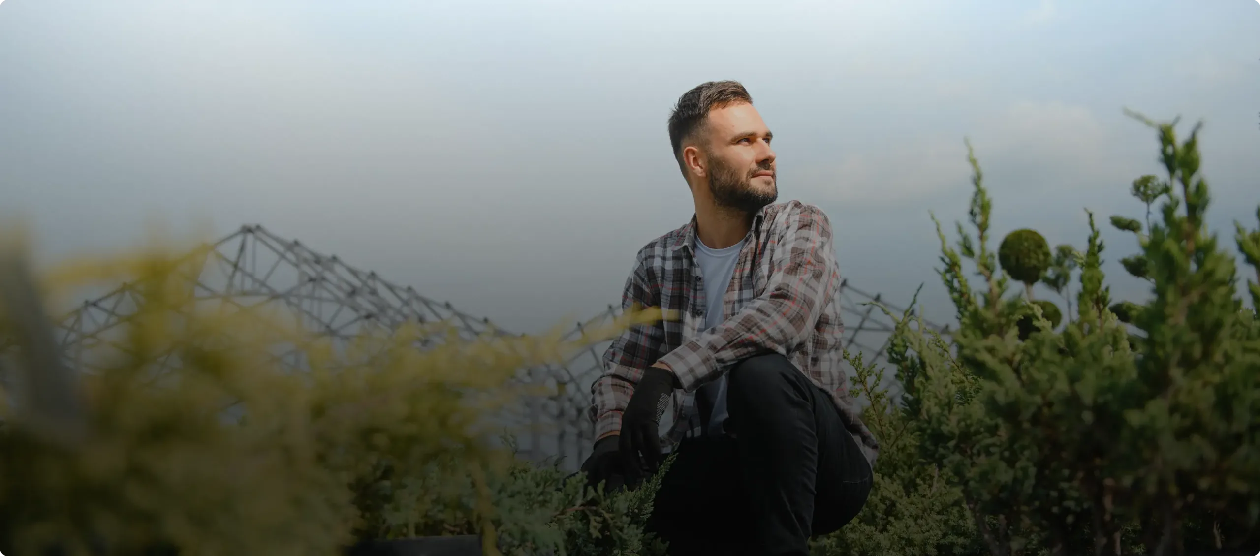 A man in a plaid shirt and gloves kneels outdoors in a garden, looking thoughtfully to the side, with greenery and a metal structure near a cannabis dispensary in the background.