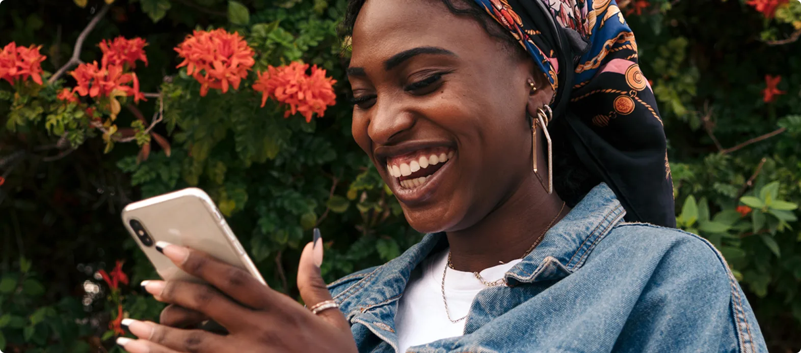 A smiling woman in a denim jacket and patterned headscarf checks her smartphone for a cannabis delivery, standing amid vibrant flowers and greenery.