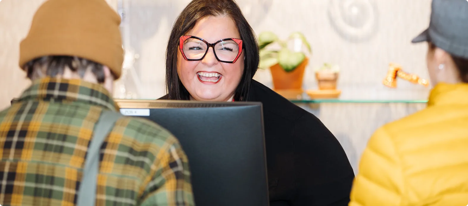 A smiling woman with large red glasses greets two customers across a cannabis dispensary counter; one wears a brown hat and plaid jacket, the other a yellow jacket.