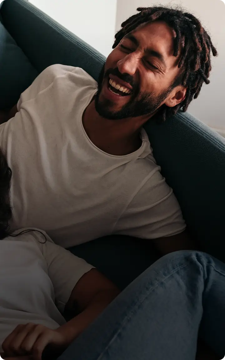 A man with short dreadlocks and a beard laughs joyfully while reclining on a couch, wearing a white t-shirt, enjoying a relaxed moment inspired by Connecticut cannabis laws. Another person is partially visible beside him.