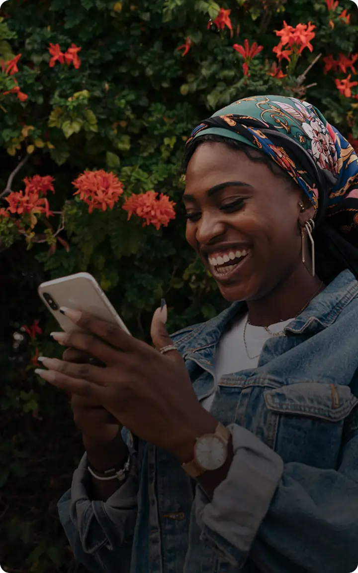 A woman in a denim jacket and colorful headscarf smiles at her phone, excitedly tracking her cannabis delivery while standing before a bush with bright red flowers.
