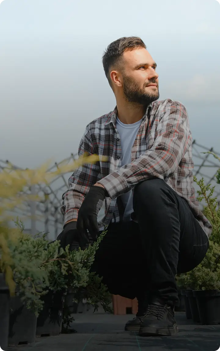 A man wearing a plaid shirt and black gloves crouches among potted plants in a cannabis dispensary greenhouse, looking off to the side and smiling.