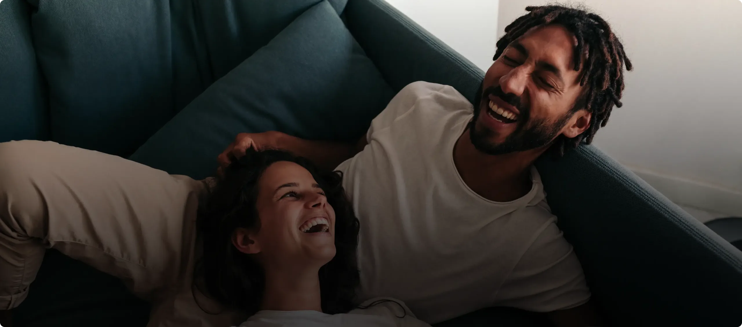 A man and woman are lying on a couch, laughing together and looking happy, with sunlight softly illuminating their cozy Connecticut cannabis moment.