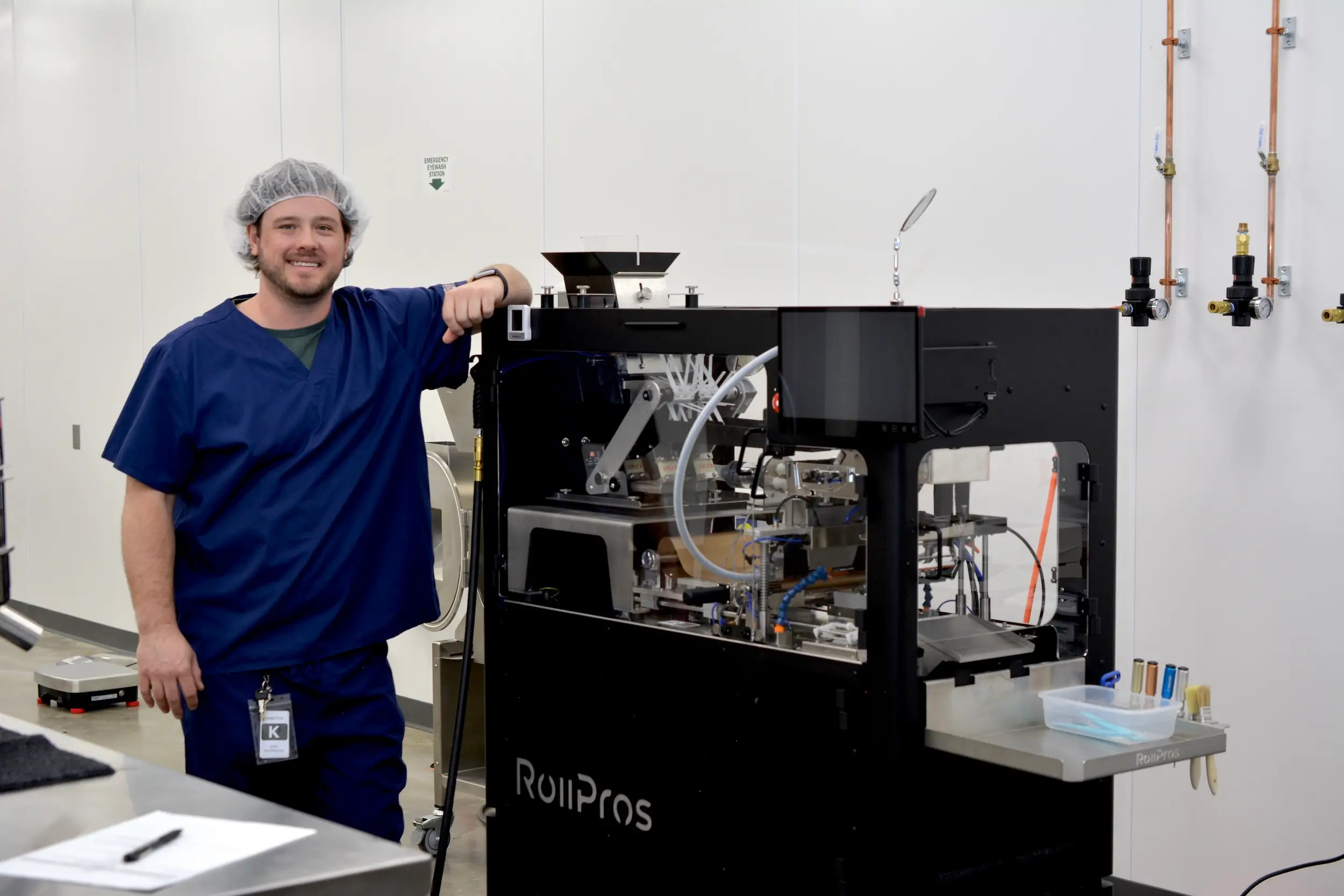 A person in blue scrubs and a hairnet stands smiling beside a RollPros machine in a clean Connecticut cannabis lab with white walls and medical equipment.