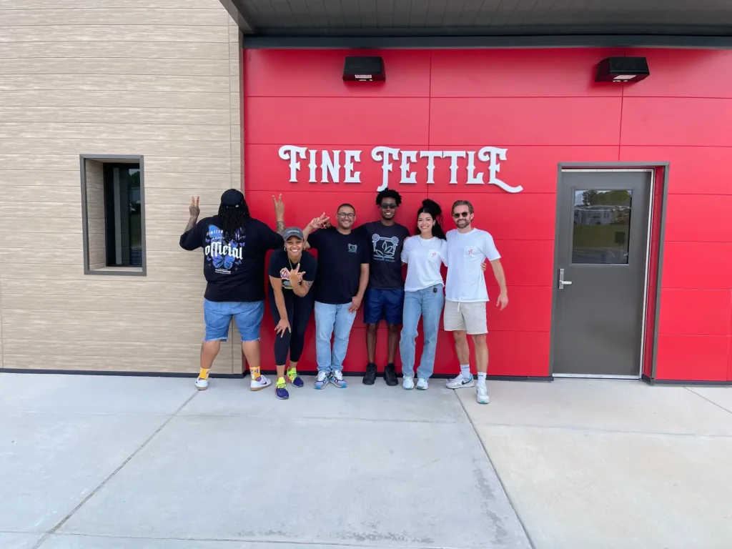 Six people stand smiling in front of a red wall with Fine Fettle written on it, posing with peace signs outside a CT cannabis dispensary. A gray door is on the right side of the wall.