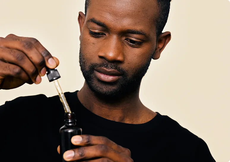 A man in a black shirt uses a dropper to take liquid from a small dark bottle, possibly exploring marijuana facts, while looking at the dropper with a neutral expression.