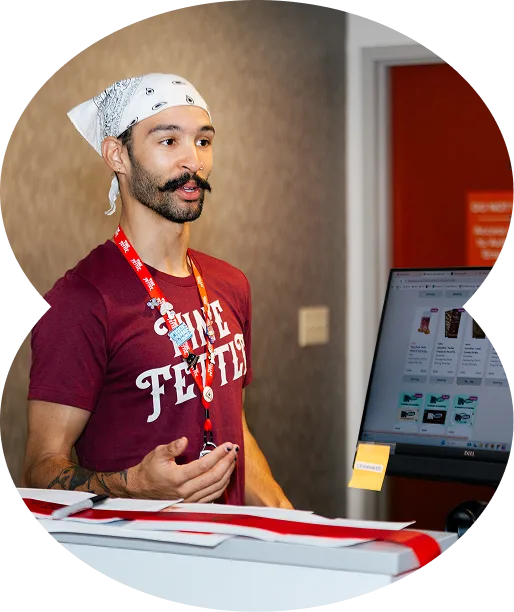 A man with a mustache and beard, wearing a bandana, maroon shirt, and lanyard, gestures while discussing the Georgia medical marijuana card at a computer desk.