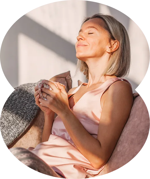 A woman with short blonde hair sits on a couch, eyes closed and smiling, enjoying a hot drink in sunlight after learning about Georgia medical marijuana.