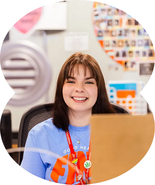 A young woman with brown hair and bangs smiles while sitting at a cannabis dispensary office desk, wearing a blue shirt and red lanyard. Colorful photos and posters brighten the space.