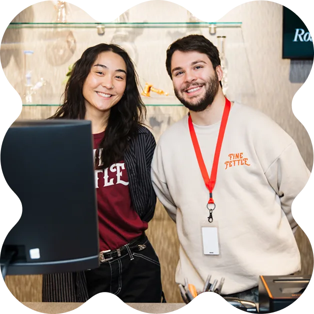 Two smiling employees at the cannabis dispensary stand behind a counter, one in a Fine Fettle sweatshirt and lanyard, the other in a maroon shirt and striped jacket, shelves behind them.