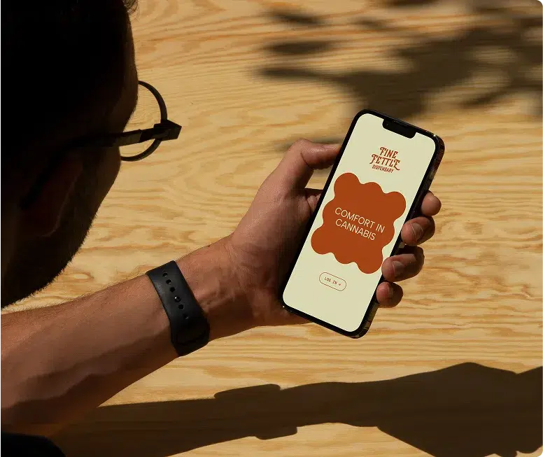 A person wearing glasses and a smartwatch holds a smartphone displaying “Comfort in Cannabis” at a light wooden table, hinting at the welcoming vibe of Rowley Dispensary.