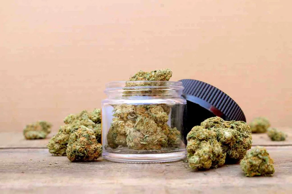 A clear jar filled with cannabis buds sits on a wooden surface, surrounded by more buds. The jar’s black lid is placed to the side.