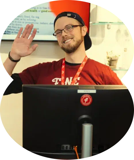 A smiling person wearing glasses and a cap waves from behind a computer monitor at a cannabis dispensary in an indoor setting.