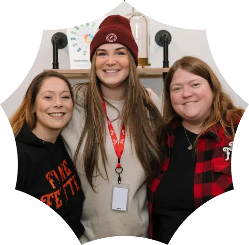 Three smiling women stand close together indoors at a cannabis dispensary; one wears a red beanie, another a red plaid shirt, and the third a black hoodie.
