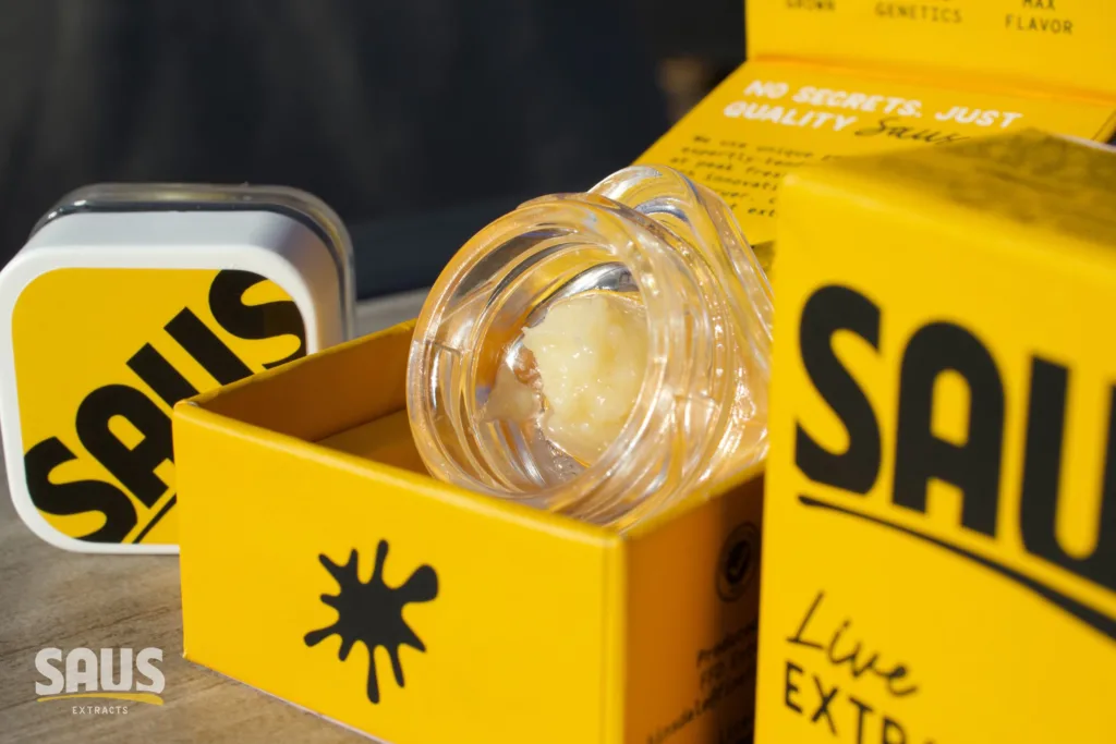 A close-up of SAUS live extracts in a clear jar, surrounded by bright yellow branded packaging and a tin with the SAUS logo.