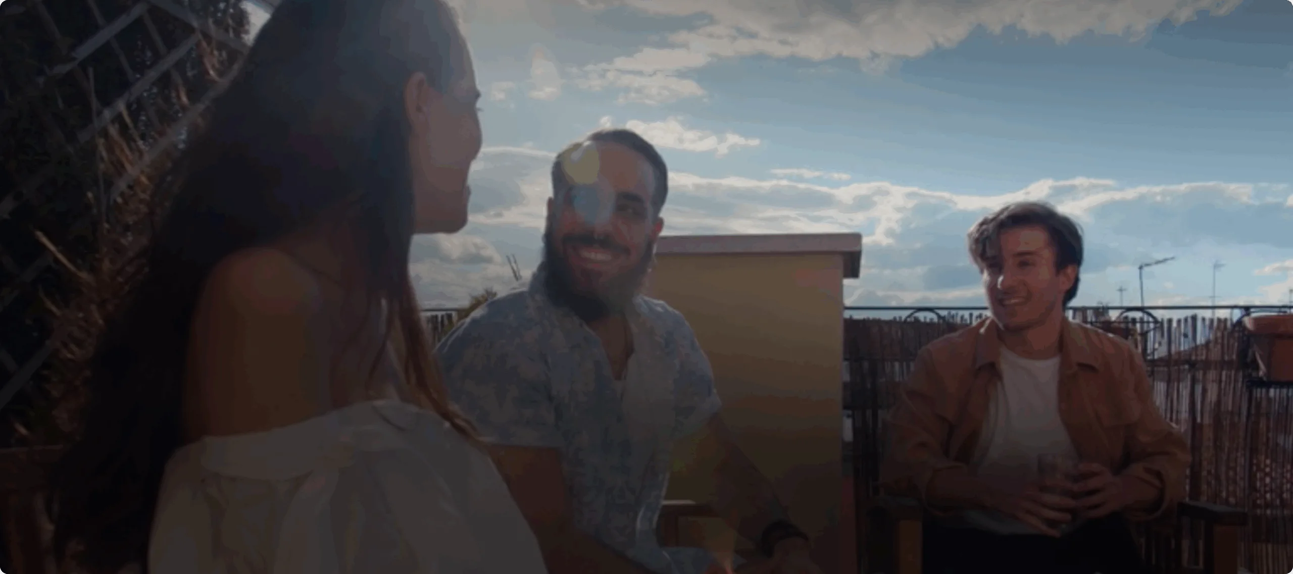 Three people sit outdoors on a sunny day, smiling and talking near a cannabis dispensary, with a blue sky and clouds in the background.