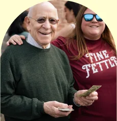 An older man smiling and holding cash stands next to a woman in sunglasses and a Fine Fettle shirt, both appearing happy outdoors at a cannabis dispensary.