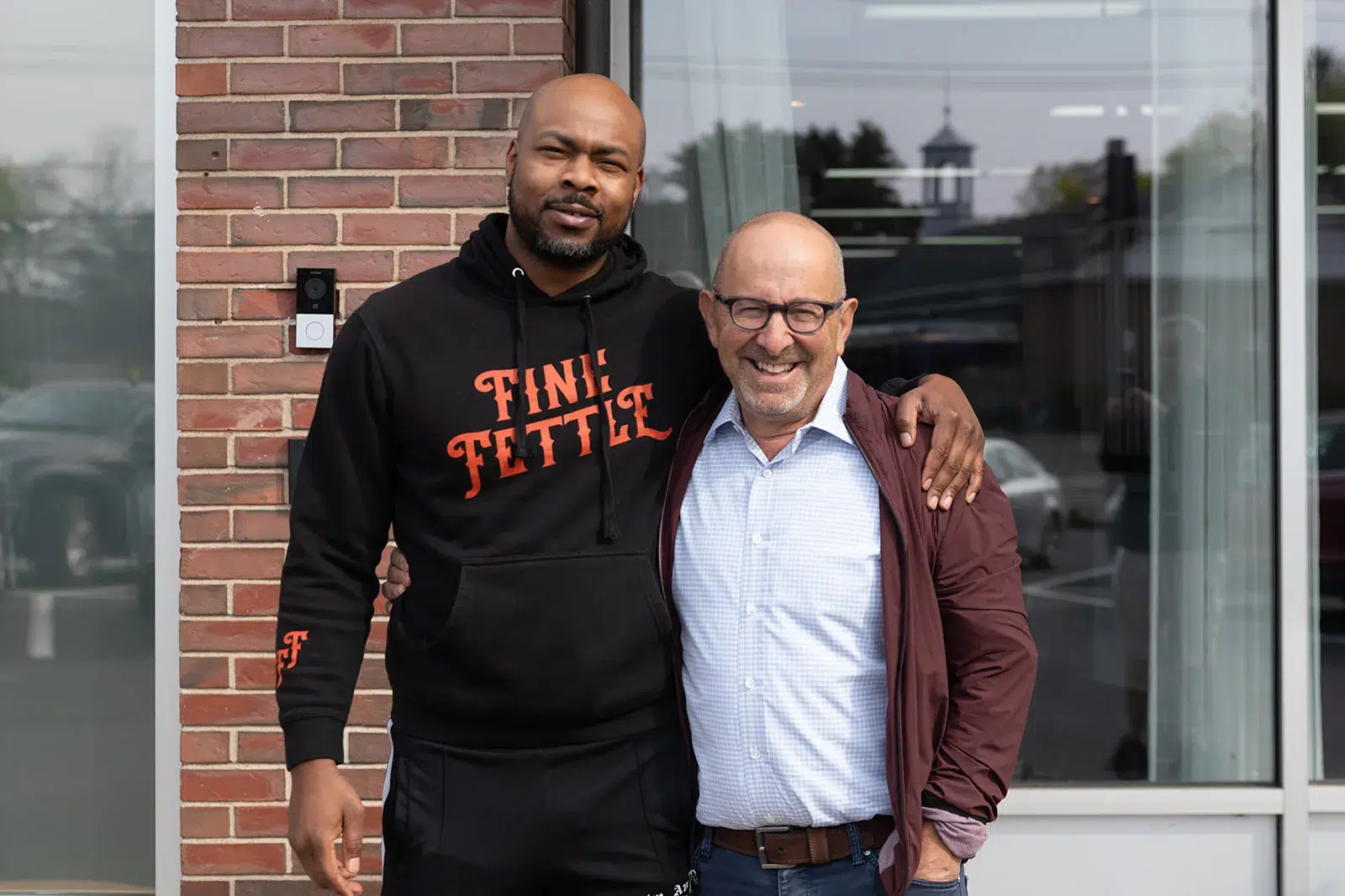 Two men stand smiling outside a building, one in a black hoodie with orange text for a cannabis delivery service, the other in glasses, blue shirt, and maroon jacket.