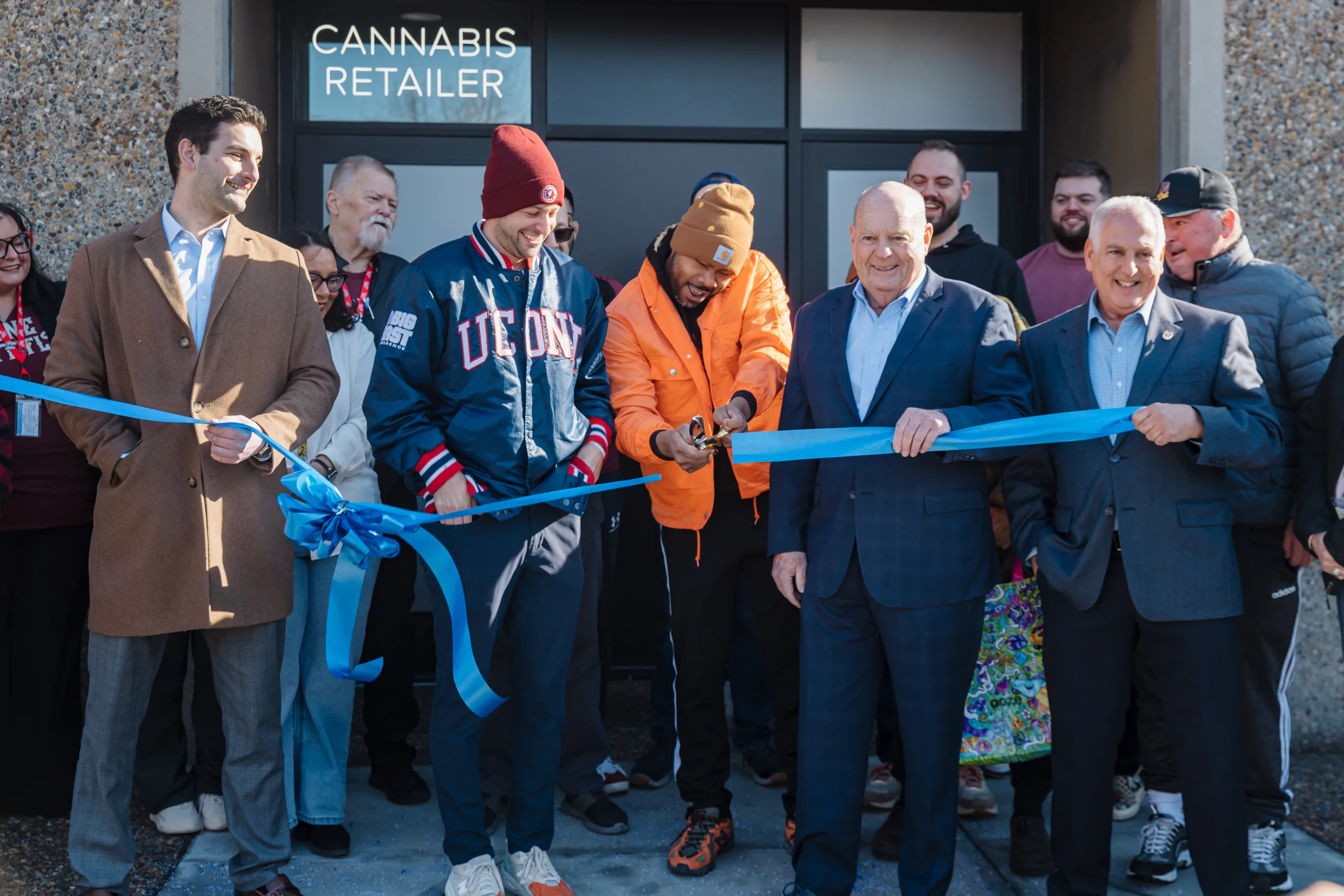 A group of people smile as one person cuts a blue ribbon in front of a building with a Cannabis Retailer sign during a ribbon-cutting ceremony.
