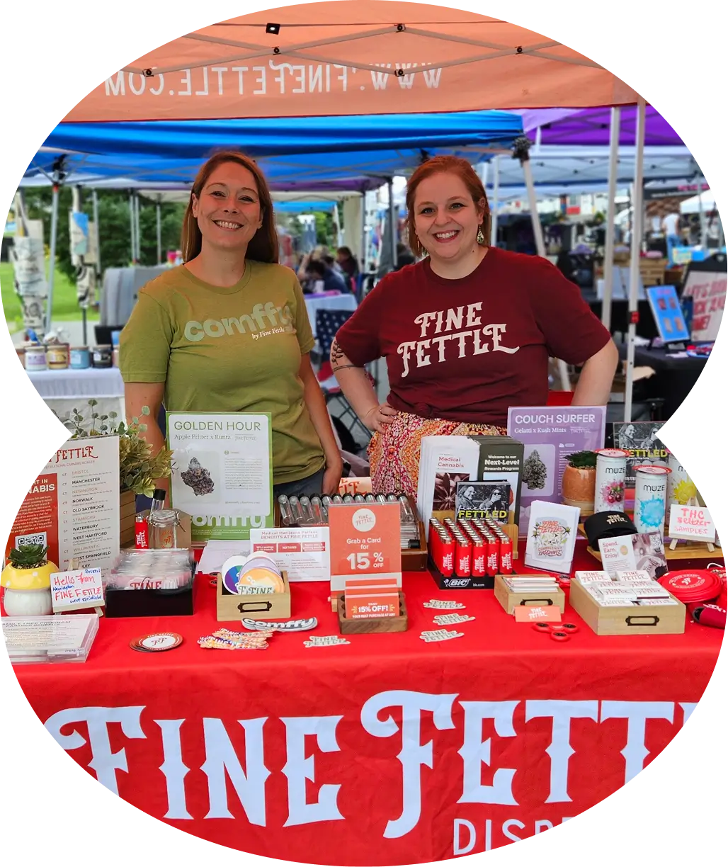 Two smiling women showcase their small business partnership agreement behind a colorful vendor booth filled with products and promotional materials under an orange tent at an outdoor market.