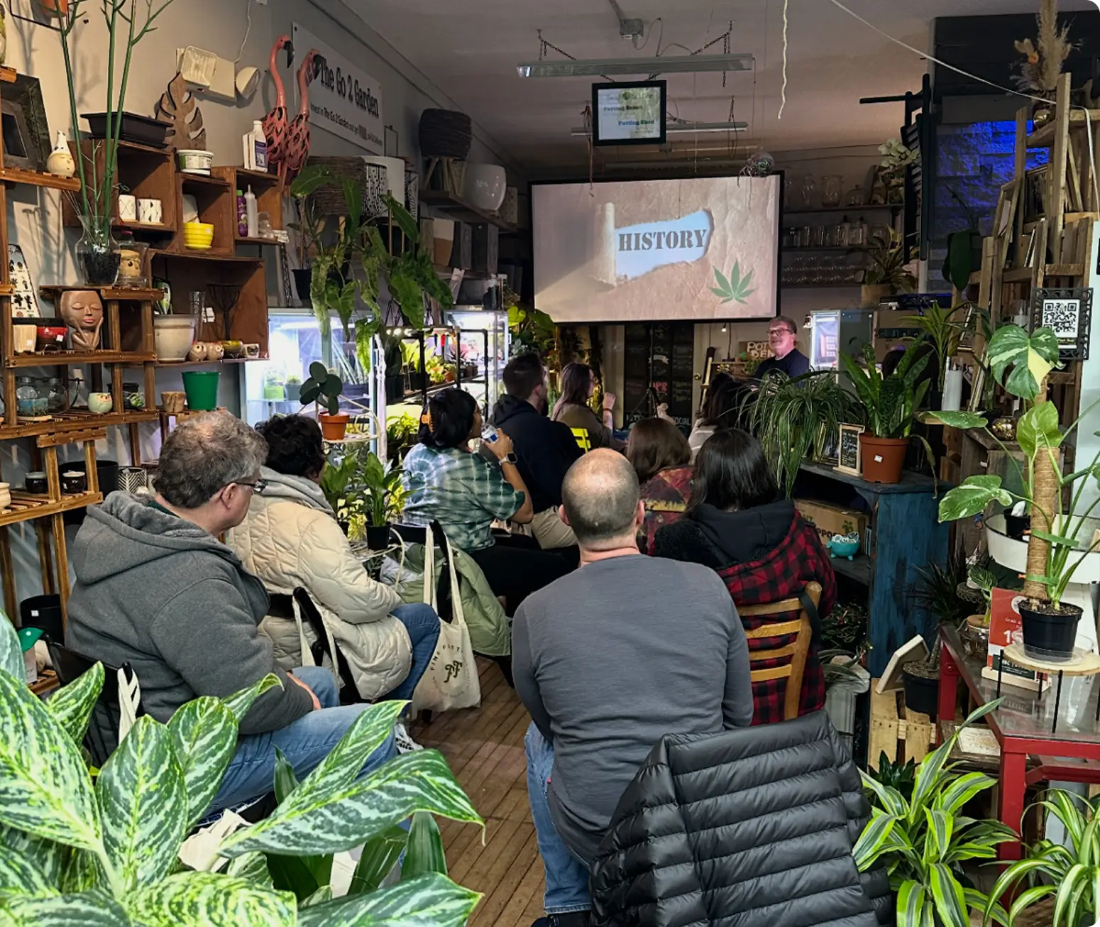 A group of people sit among potted plants in a cozy room, watching a presentation on HISTORY with a cannabis leaf, discussing a small business partnership agreement.