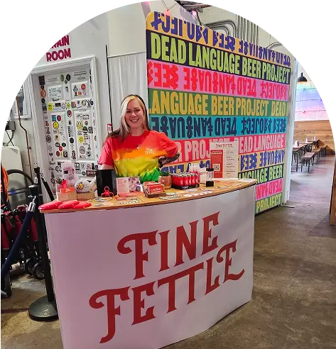 A smiling woman stands behind a Fine Fettle booth with colorful products, highlighting a small business partnership agreement at a Dead Language Beer Project event indoors.