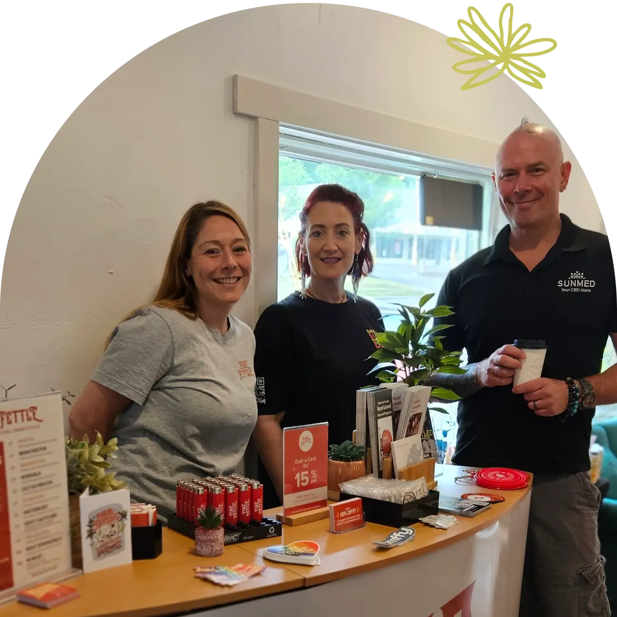 Three people stand and smile behind a wooden counter with small plants, flyers, and promotional materials for their small business partnership agreement in a well-lit indoor setting.