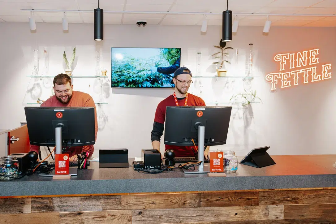 Two smiling employees stand behind a wooden counter at Fine Fettle, working at registers. Discover our welcoming dispensary locations with plants and a screen in the background.