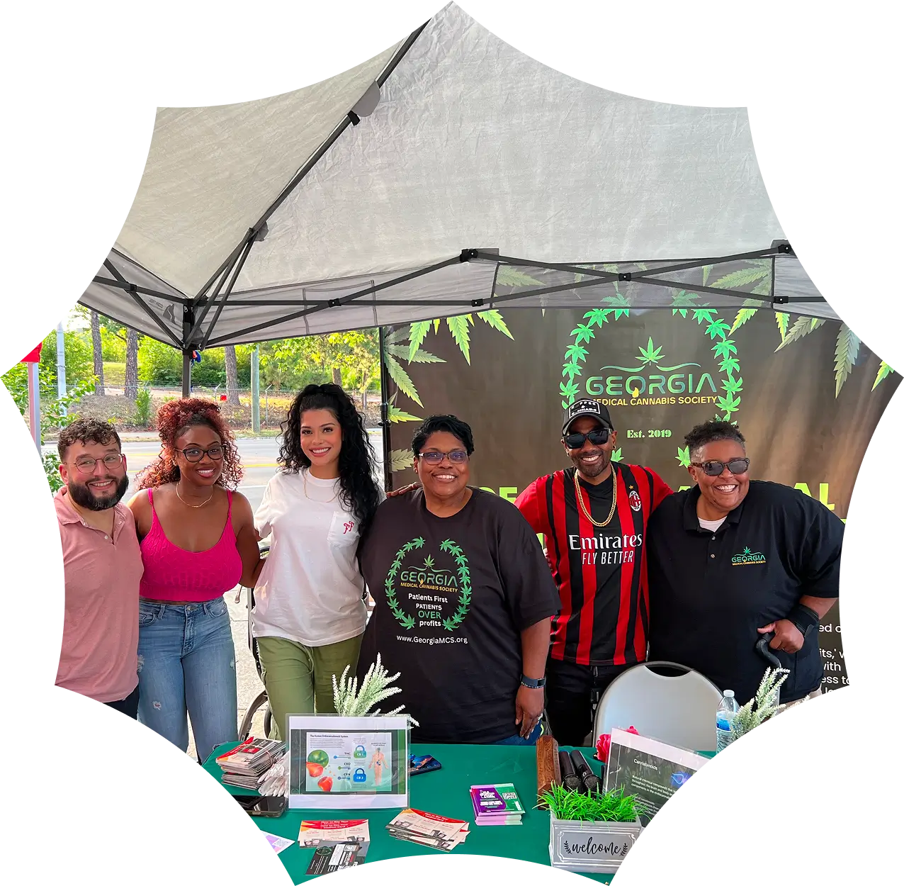 Six people smile together under a canopy at a booth for the Georgia Cannabis Society, with informational materials and banners displayed on the table.