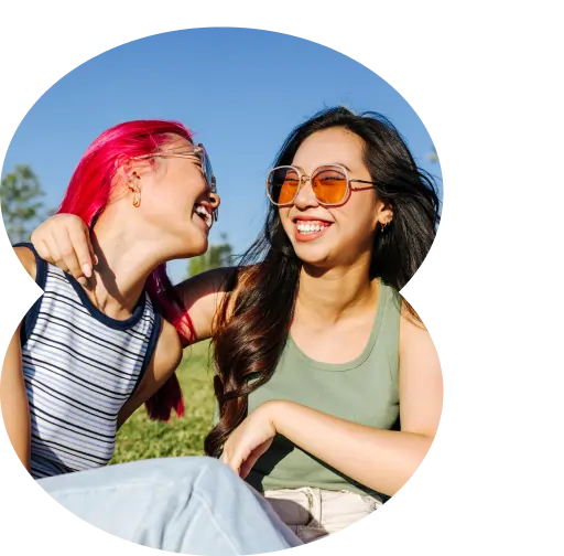 Two young women sitting outdoors on grass, smiling and laughing while sharing fun marijuana facts. One has bright pink hair, the other has long dark hair and sunglasses, blue sky behind them.