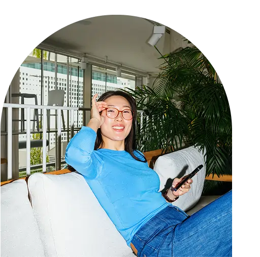 Woman sitting on a white couch, smiling, holding a phone, and making an OK gesture with her hand; large windows and plants in the background as she reads marijuana facts.