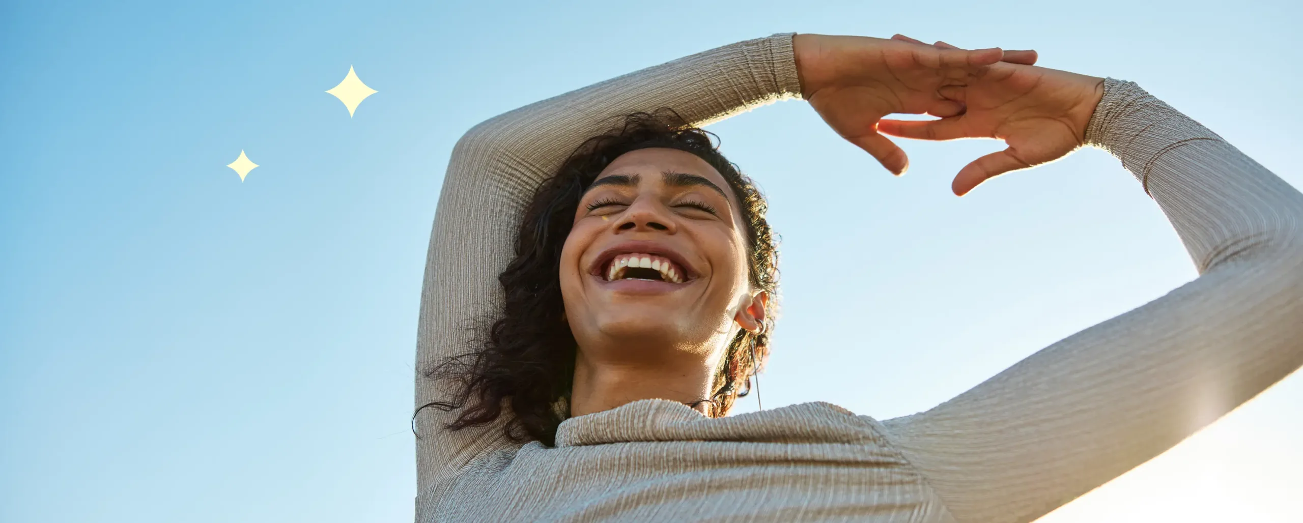 A woman smiles joyfully with her arms raised against a clear blue sky, sunlight illuminating her face. A yellow star shape appears in the background.