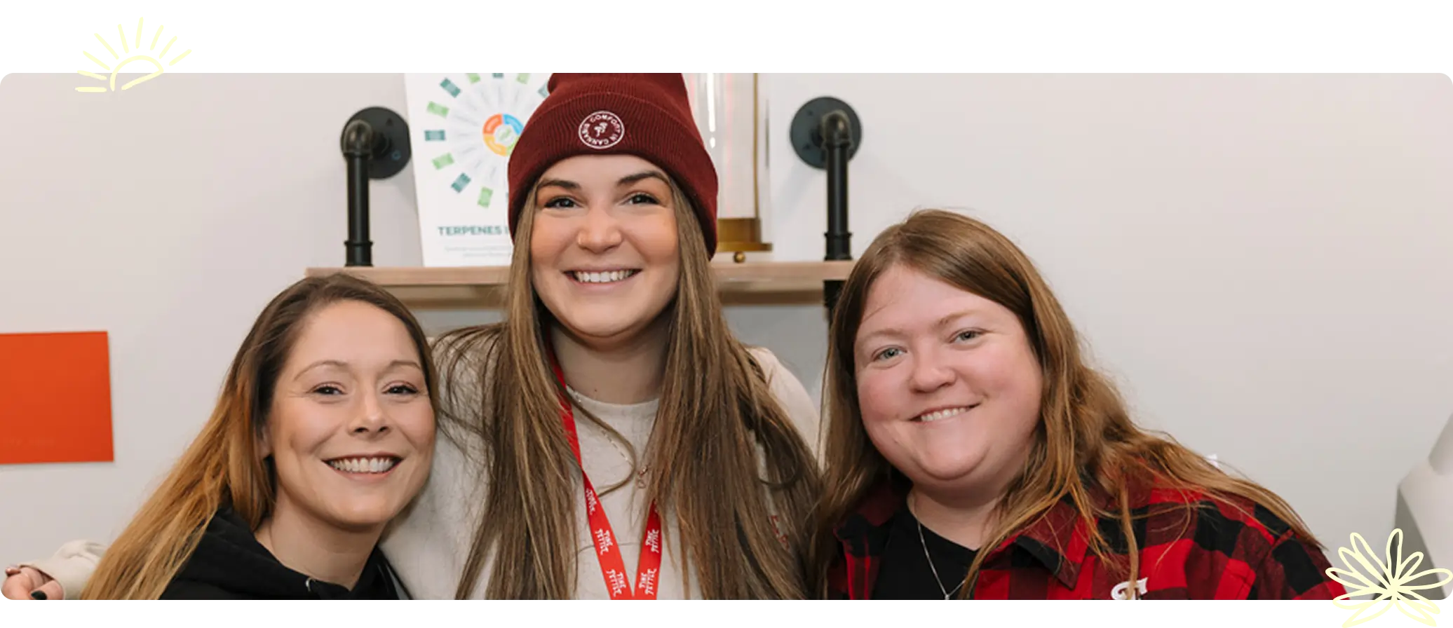 Three women stand close together smiling at the camera, one wearing a maroon beanie and lanyard, in a casual cannabis dispensary setting.