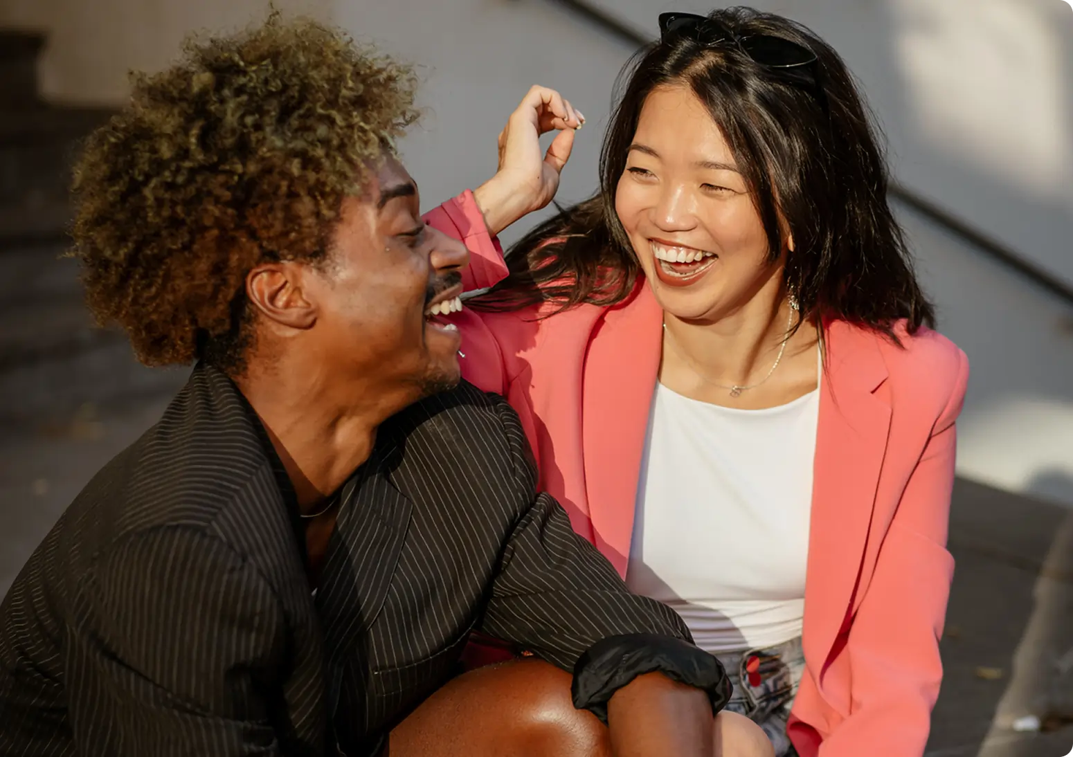 Two friends sit on outdoor steps, dressed in blazers, laughing and sharing marijuana facts with each other in the bright sunlight.