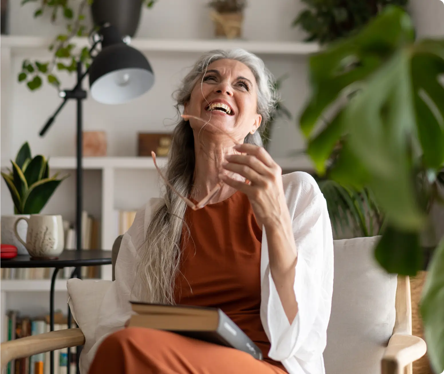 Older woman with long gray hair sits in a cozy living room, smiling and laughing, holding glasses and a book, chatting about local events amid plants and bookshelves.