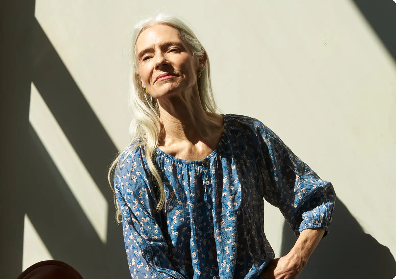 An older woman with long white hair, wearing a blue floral blouse, stands in sunlight against a plain wall, looking content and relaxed as she reads about marijuana facts.