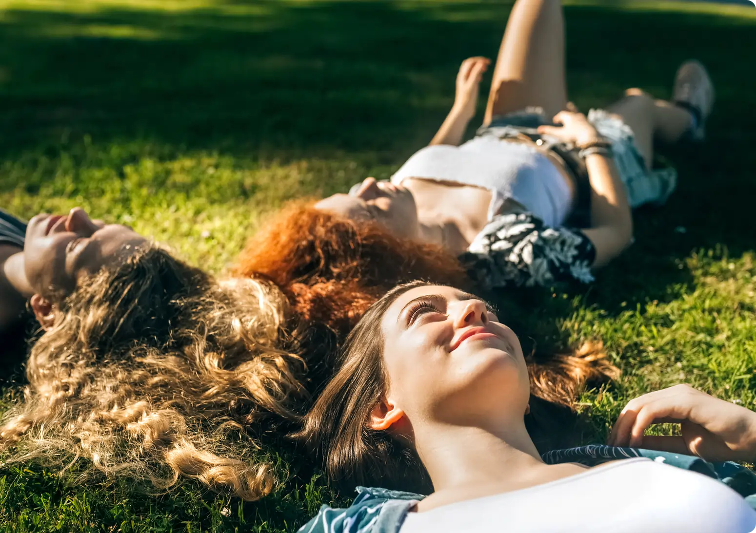 Three young women lying on grass in a park, looking relaxed and happy as they enjoy the sunlight and discuss interesting marijuana facts.